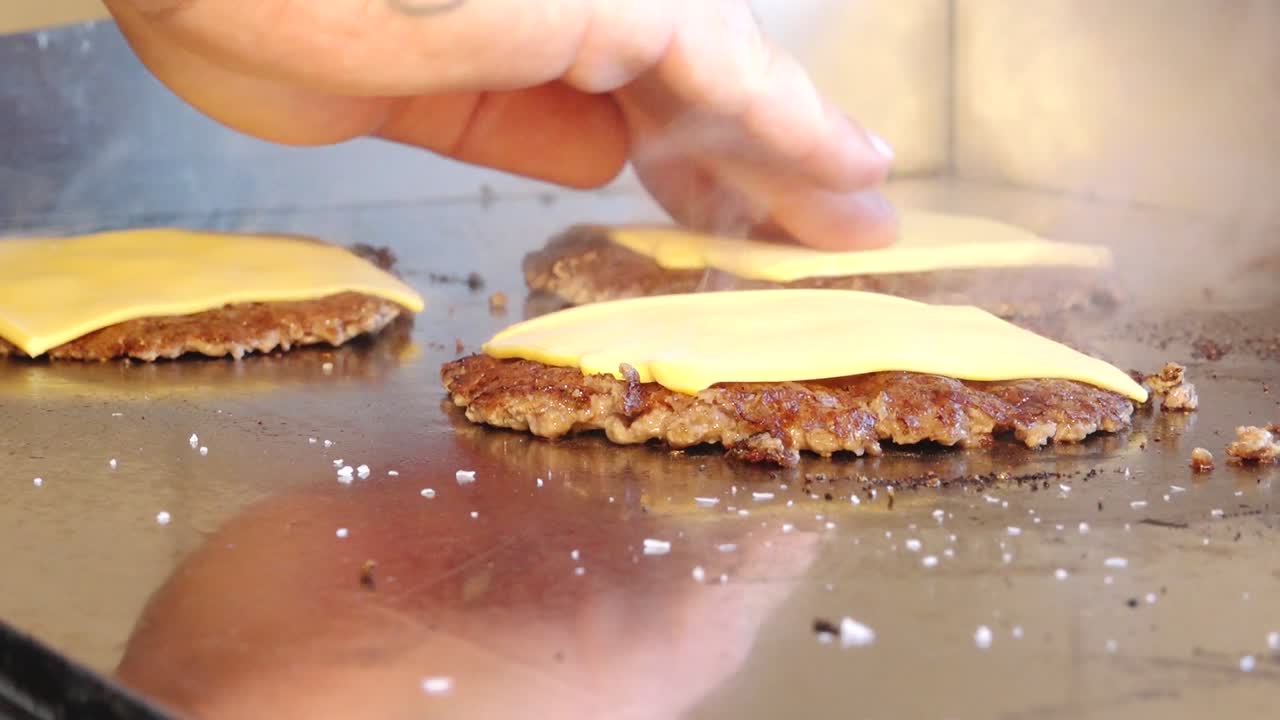 Primo piano di hamburger di manzo che cuociono su una piastra calda, ricoperti da fette di formaggio fuso. Il vapore sale dalla superficie, evidenziando la consistenza e i dettagli succosi