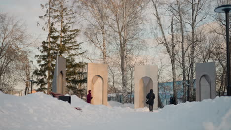 winter season scene showing people walking on snow covered path near tall sculptural monuments, with one person in red hoodie running and another in black coat walking in center under soft daylight