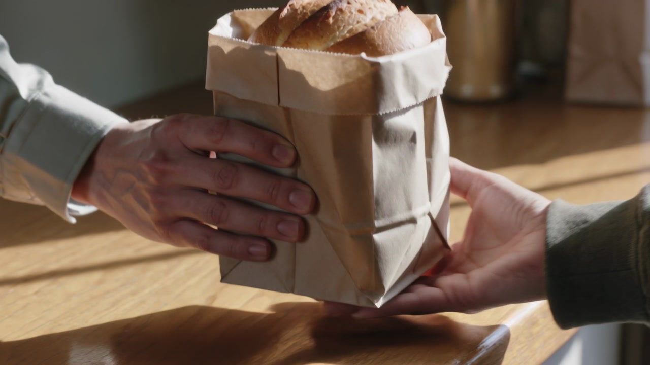 Placing paper bag, seller wearing shirt, dark apron handing loaf to buyer finishing sale at counter