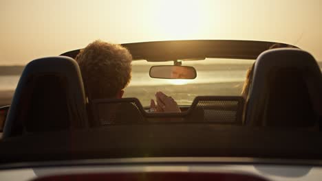 A-guy-and-his-girlfriend-are-talking-in-a-convertible-car.-A-girl-and-a-boy-sit-in-a-convertible-car-and-hold-each-other's-hand-in-windy-weather-against-the-river-and-a-yellow-sky