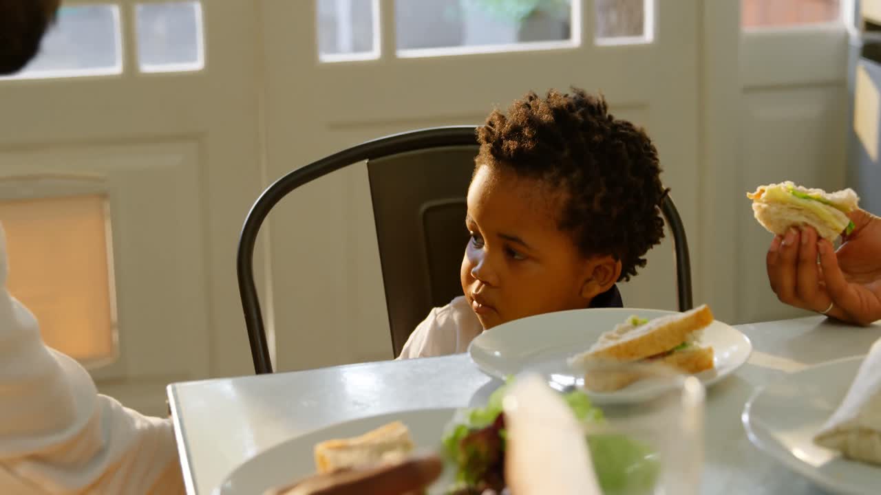 Front view of cute little black son eating food at dinning table in kitchen of comfortable home 4k