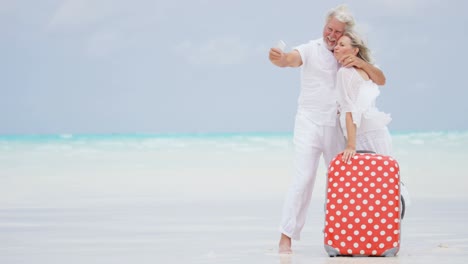 caucasian seniors taking selfie on beach with suitcase