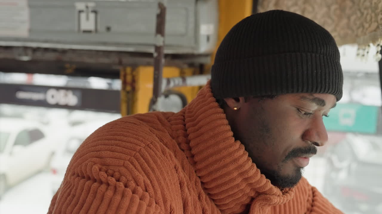 Black man steering bus through traffic, focused expression, hands on wheel, snowy urban scene outside, driver checking mirrors, rush hour tension, professional shift mood, attentive navigation