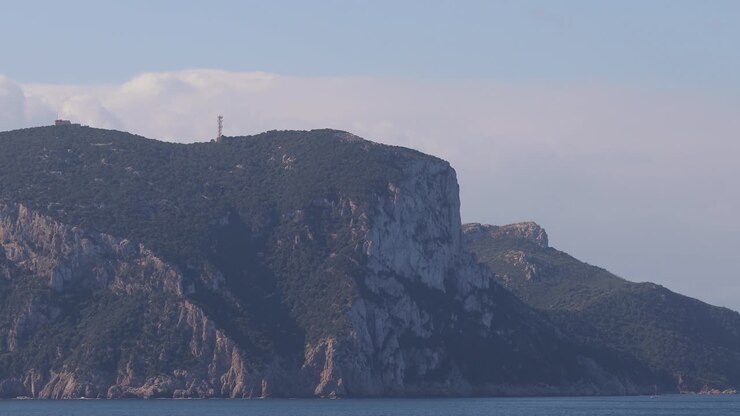 Isola con scogliere imponenti vista dal mare aperto e blu, sotto un cielo nuvoloso e pallido.