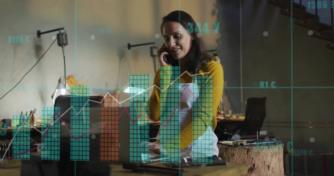Woman holding phone to ear and typing on laptop in workshop, analyzing evolving business bar charts