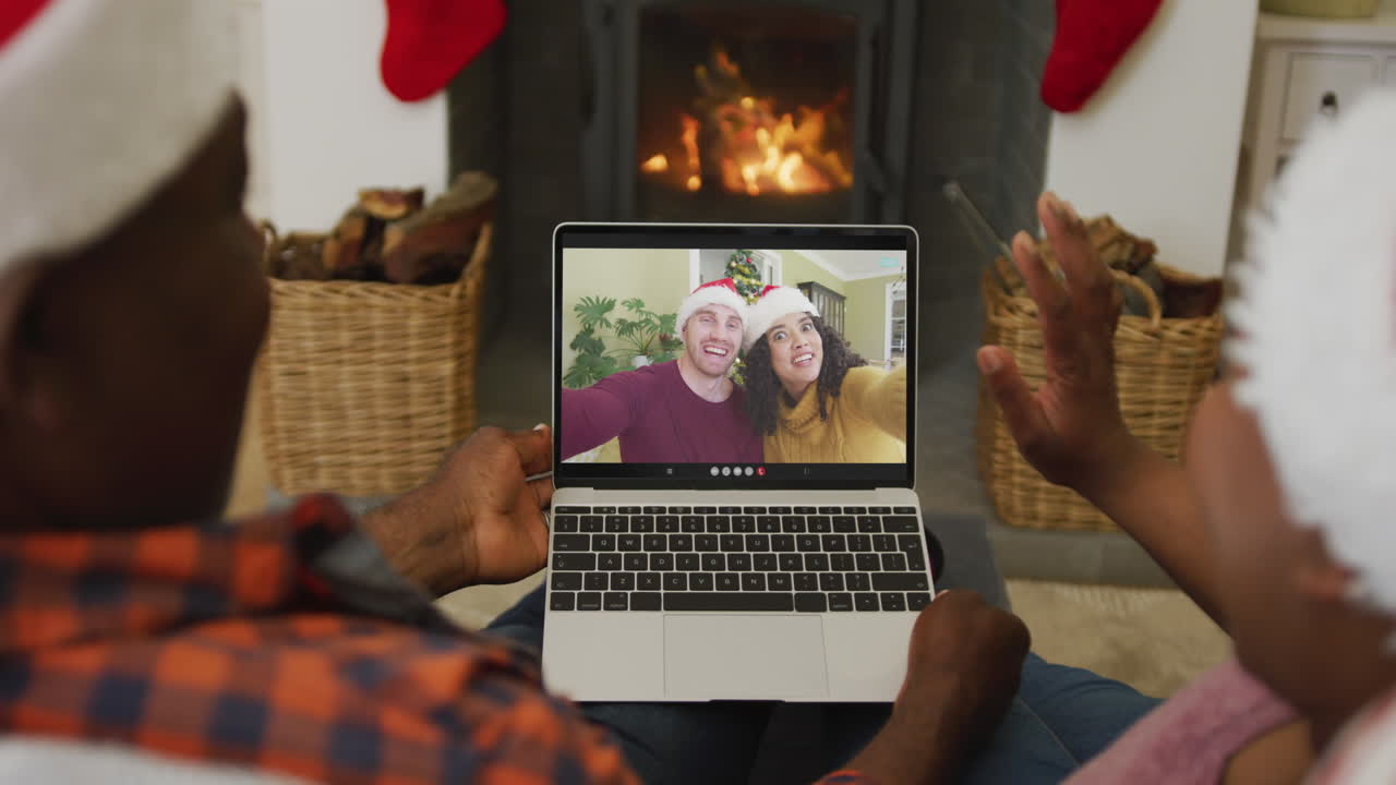 pareja afroamericana con sombreros de santa usando una computadora portátil para una videollamada de navidad con la pareja en la pantalla