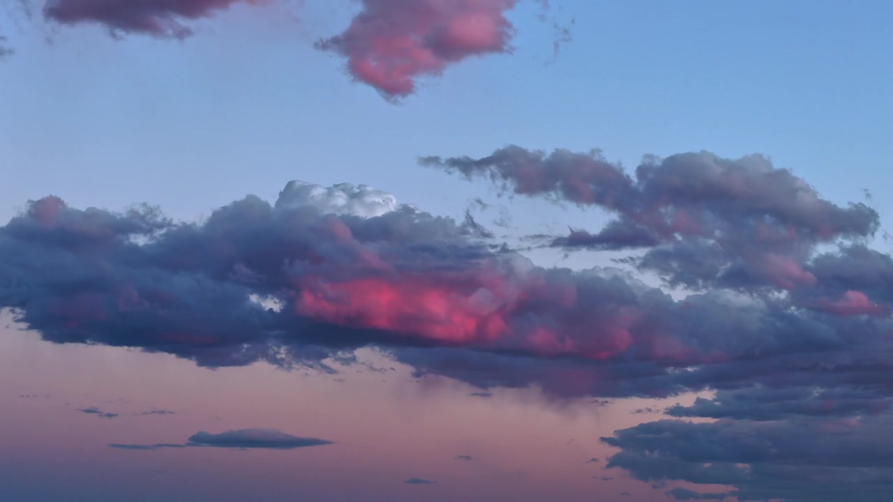 4K drone timelapse focused on a cluster of twilight clouds. A striking, neon magenta sunset glow illuminates the textured center cloud before slowly fading away into the darkening blue evening sky.