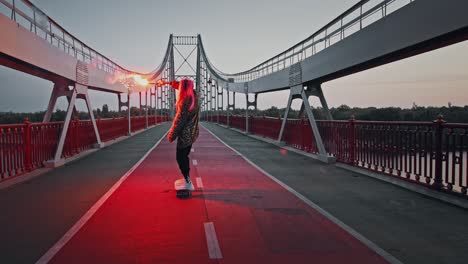 young hipster woman in informal outfit is riding skateboard on bridge holding burning red signal flare and waving it