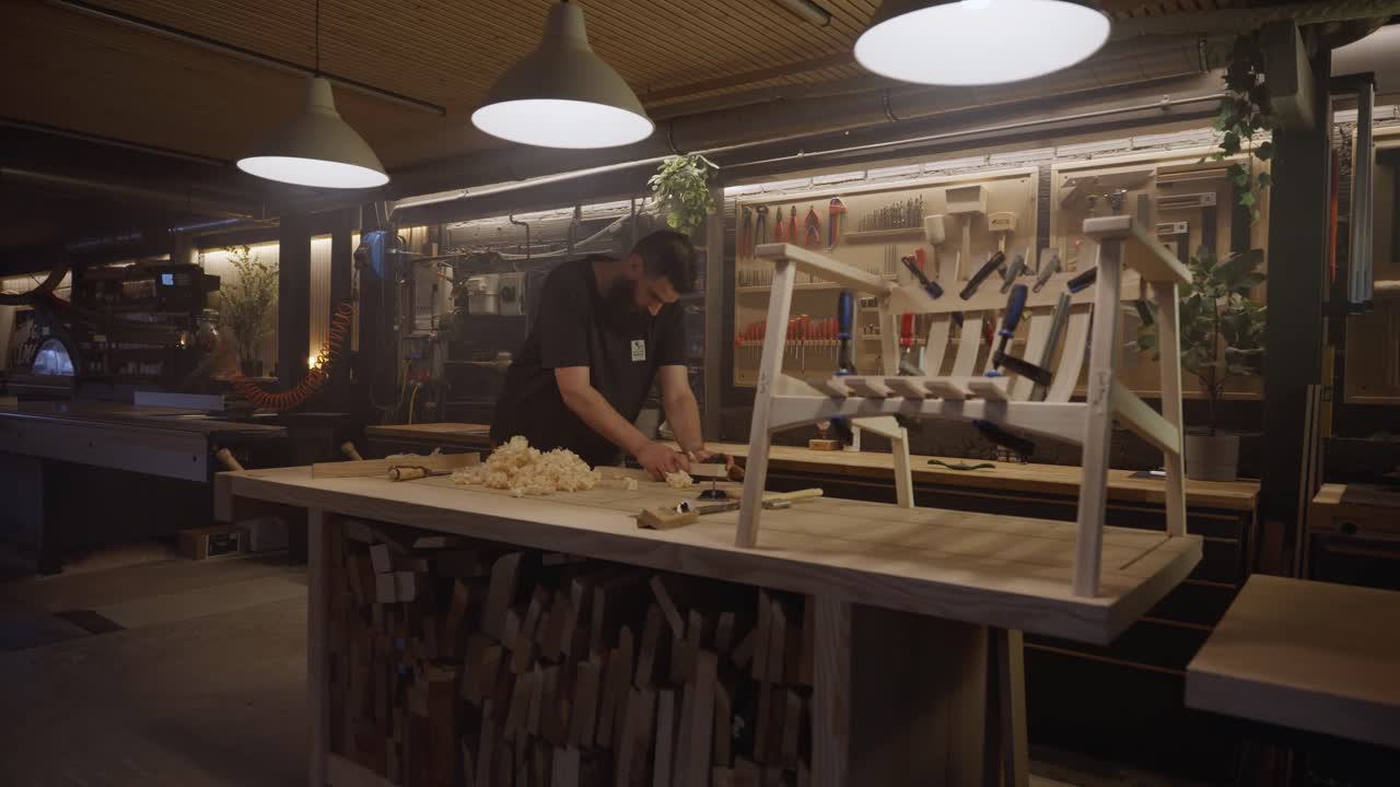 A craftsman stands in a modern woodworking studio surrounded by tools, a workbench, and a wooden furniture frame, representing craftsmanship, creativity, and handmade design