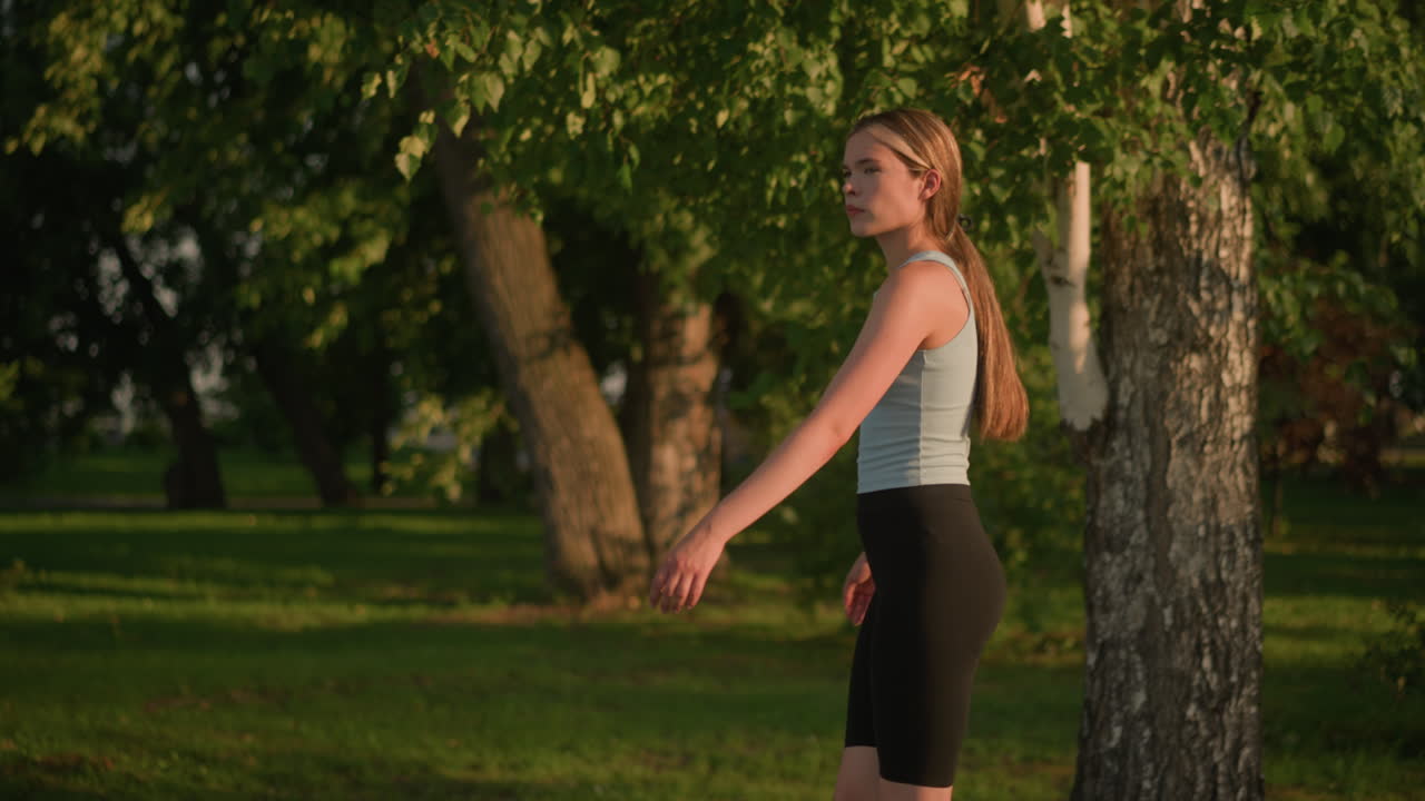 vista lateral de una joven que parece infeliz mientras se mueve con gracia como patinaje sobre ruedas al aire libre, la luz del sol se refleja en ella, el cabello atado hacia atrás, fondo natural con árboles verdes exuberantes
