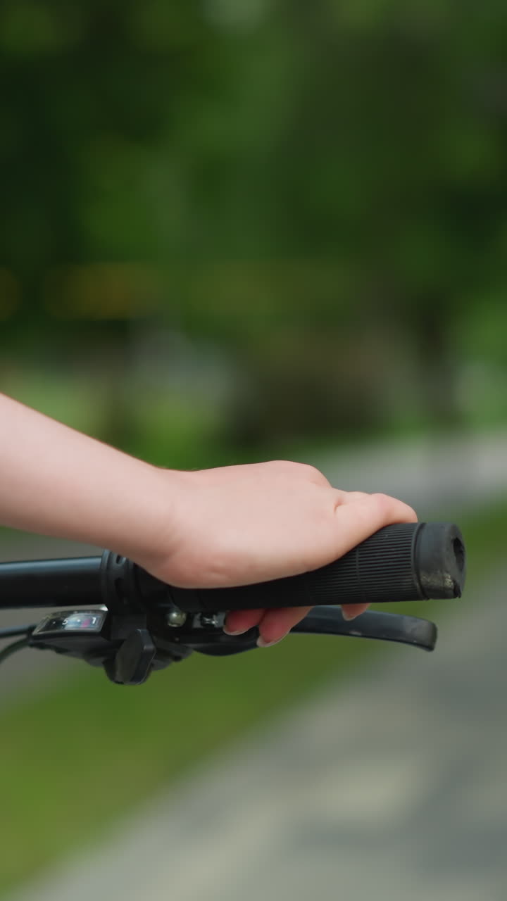 primer plano de la mano de la persona agarrando el manillar de la bicicleta mientras se pasea por un camino del parque, rodeado de vegetación borrosa y una figura fuera de foco en el fondo