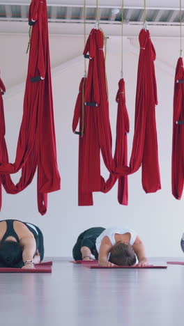 relaxed ladies sit in extended child poses under hanging long red fly yoga hammocks in modern gym stretching zone