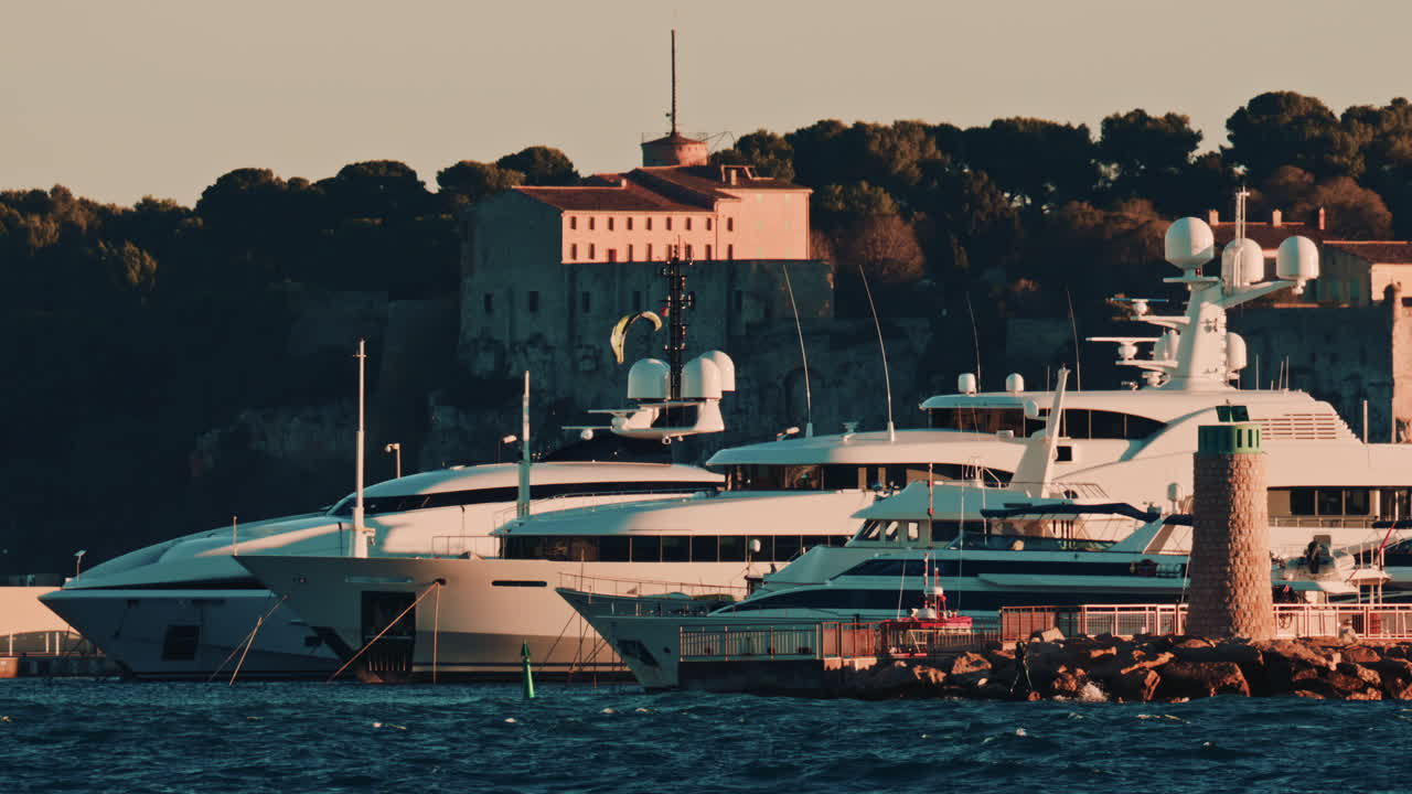 Luxury yachts docked near a stone lighthouse, with a historic building and trees in the background, port of Cannes, France