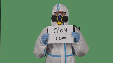 portrait of a man in a protective suit, goggles, gloves and a respirator looking at the camera holding a stay at home sign. green screen, chroma key. slow motion. close up