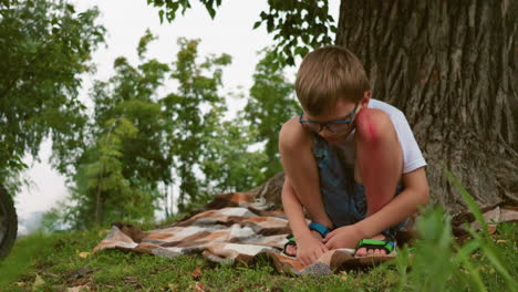 a young child sits under a tree, deep in thought, with a noticeable bruise on his leg, his hands rest on the ground, and a blanket lies beneath him