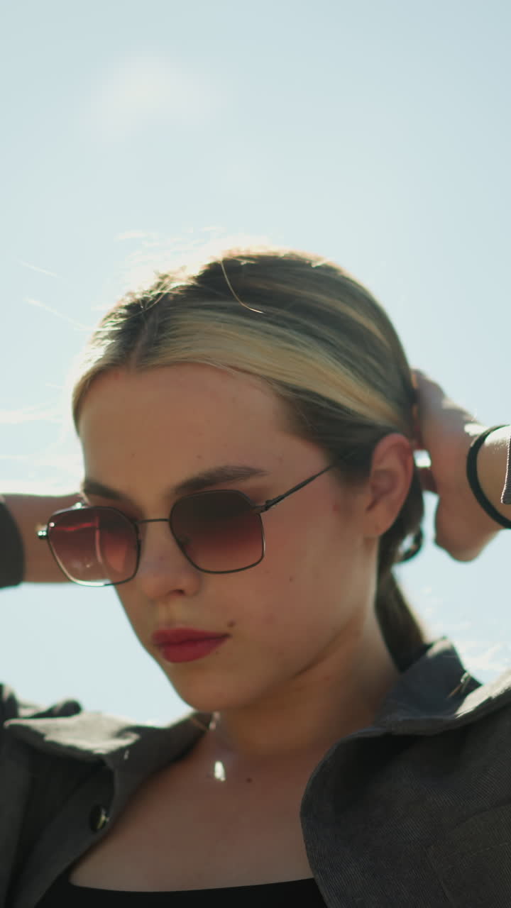 회색 재 에 검은 을 입은 여성, 밝은 빛 하늘 아래 집중된 표정으로 머리를 조정, 그녀는 선글라스를 착용, 그녀는 야외에 서