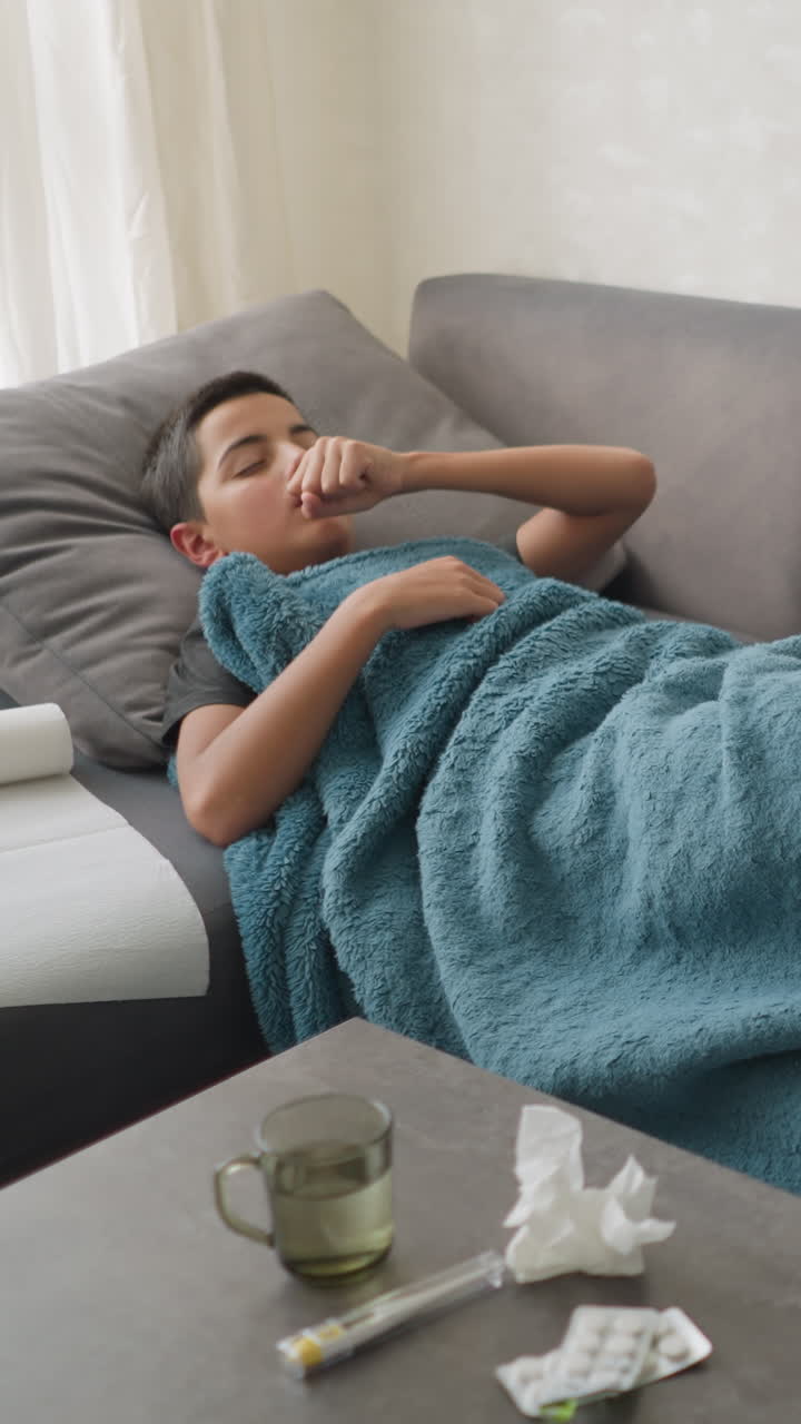 Scattered room with boy lying on couch under blanket, adjusting while glass cup and tissue papers are on the table, messy environment showcasing a sick child recovering at home