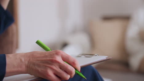 lower view of mindful observer seated in calm setting writing with green pen on notebook during thoughtful session, showing blue sleeve and partial hand with blurred reflection in background