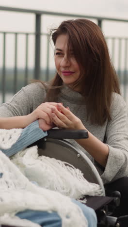 happy mother holds hand of little daughter in wheelchair on viewpoint overlooking river. young woman talks to girl with disability squatting nearby