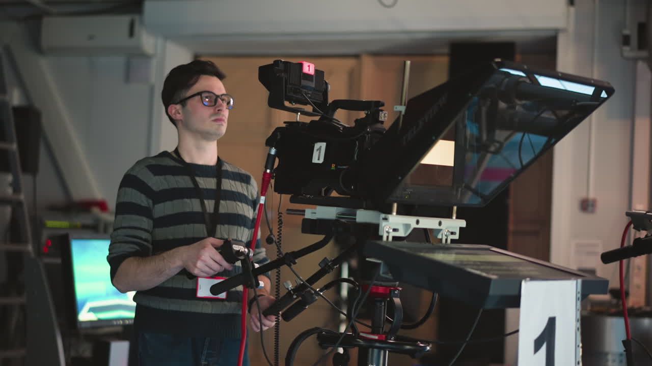 Man operating professional TV camera with teleprompter in broadcast studio. Monitor shows studio backdrop. Operator wearing striped sweater and glasses, engaged in filming process