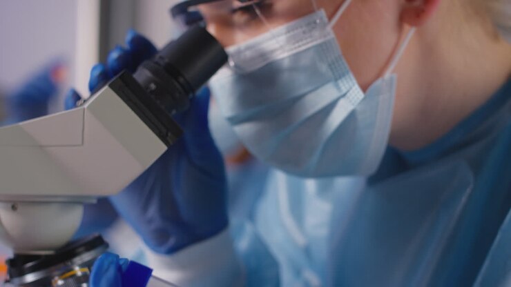 Female Lab Worker Wearing PPE Analysing Samples With Microscope Holding Test Tube Labelled Omicron