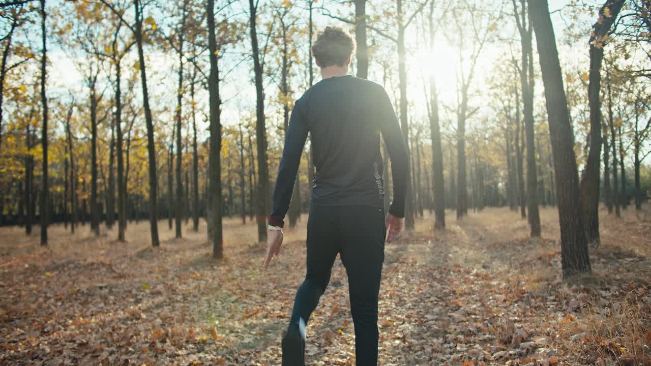 vista traseira de um homem com cabelos encaracolados em um uniforme esportivo preto aquecendo-se antes de começar sua corrida de manhã na floresta de outono. hábitos saudáveis e estilo de vida saudável correndo de manhã