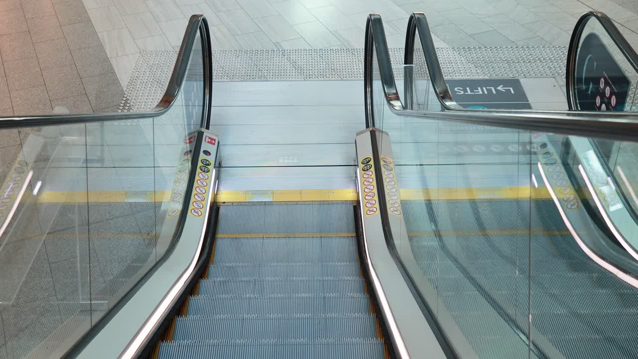 A stationary camera captures a clean, empty escalator moving downward in a brightly lit, tiled indoor environment with glass railings and clear signage