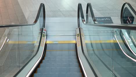 a stationary camera captures a clean, empty escalator moving downward in a brightly lit, tiled indoor environment with glass railings and clear signage