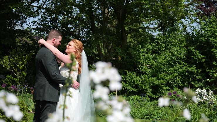 Caucasian bride and groom hold each other in garden, seen from behind flowers
