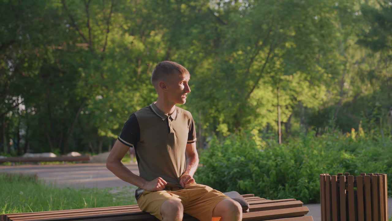 Uneasy Man Adjusting Strap In Park Ambiance, Restless Man Shifting And Fidgeting On Wooden Park Bench, Fidgeting Individual Exhibiting Nervousness While Sitting On Leafy Park Bench