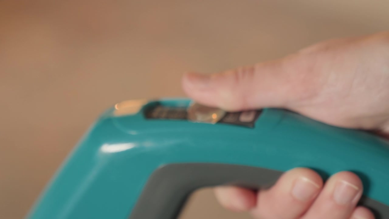 家の掃除中、クローズアップされた手が掃除機のスイッチを調整している。集中した家主がティール色のハンディ掃除機のコントロールとスライダーを確認。ゆっくりとした意図的な指の動きが、設定と握りの詳細を示している。