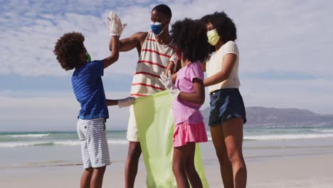 african american parents with two children wearing face masks collecting rubbish from the beach