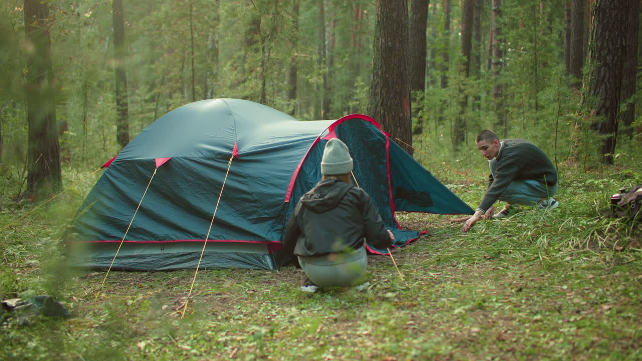 Campers focused on securing tent in forest as woman squats near entrance holding rope while man crouches nearby adjusting peg surrounded by trees and soft daylight