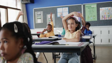 Happy-diverse-schoolchildren-at-desks-raising-hands-in-elementary-school-classroom-in-slow-motion