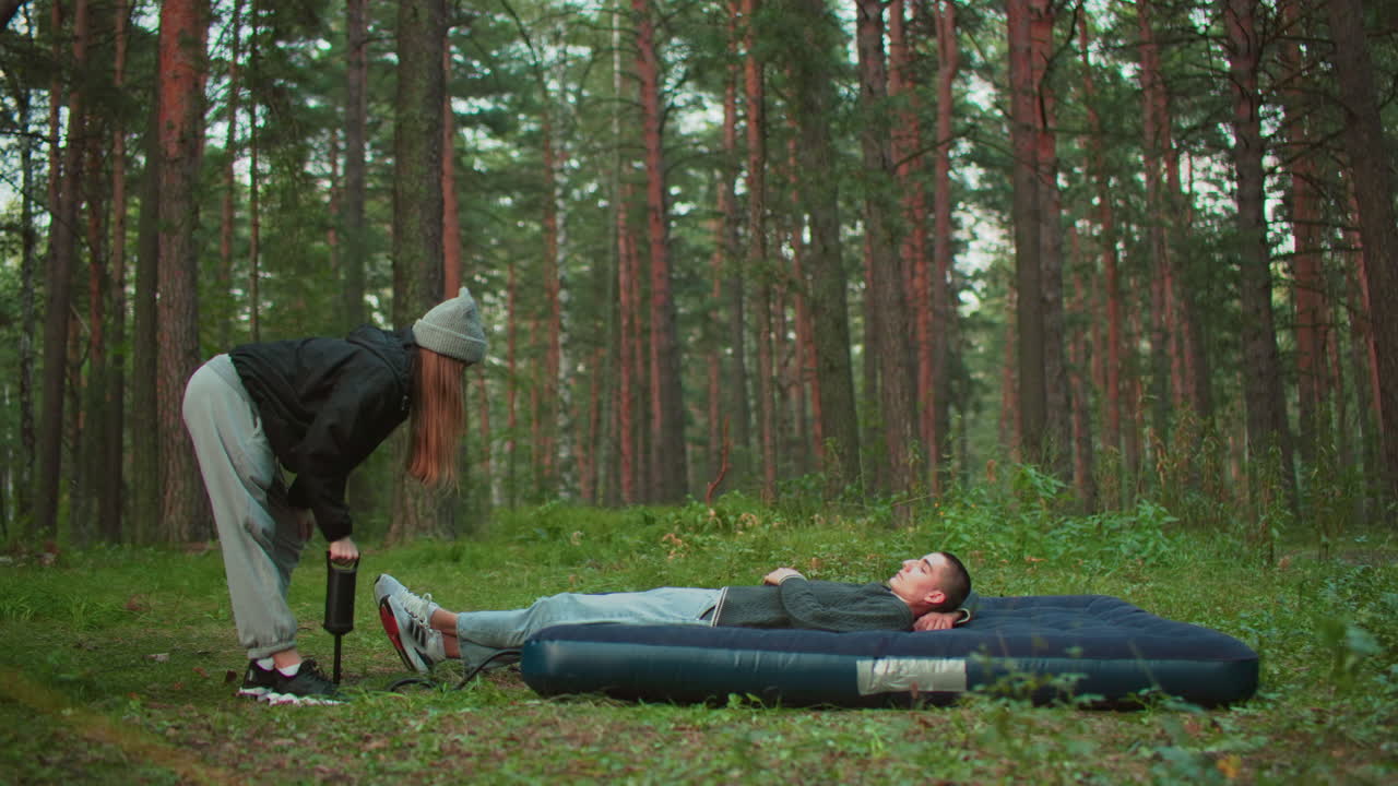 man relaxes on air bed while woman bends over pumping air into it using hand pump in forest setting, surrounded by tall trees and greenery