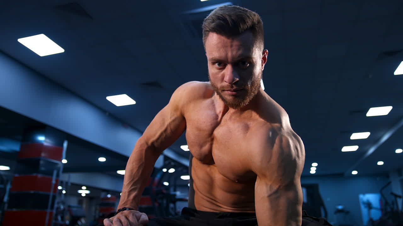 Handsome strong young bodybuilder work out with dumbbells. Strong abs. Modern gym background. Video from below.