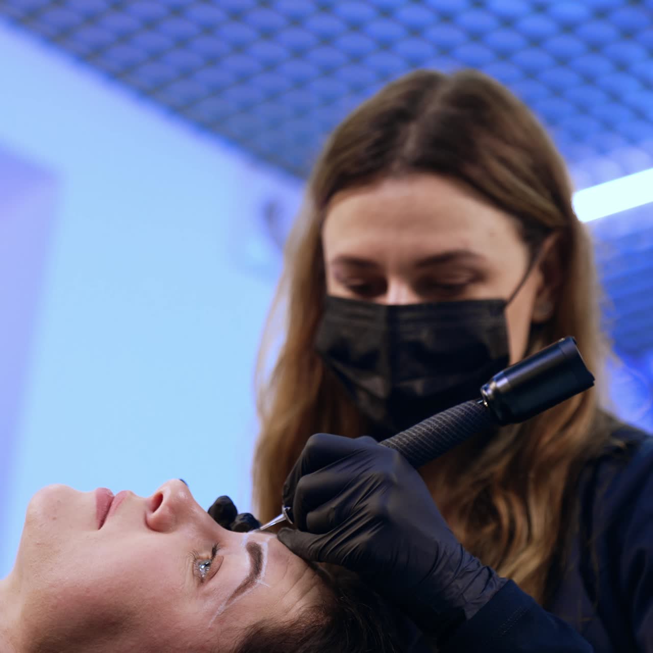 Beautician in mask and gloves performs eyebrow make up. Brunette woman lies on the couch having permanent on her brows