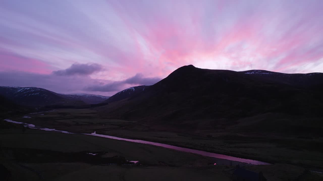 Aerial drone footage rising slowly in Glenshee, Scotland during an intense pink and purple sunset looking towards the silhouetted mountains as the sunset reflects in a river and off of the clouds