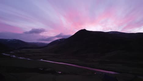 Imágenes-Aéreas-De-Drones-Que-Se-Elevan-Lentamente-En-Glenshee,-Escocia,-Durante-Una-Intensa-Puesta-De-Sol-Rosa-Y-Púrpura-Mirando-Hacia-Las-Montañas-Silueteadas-Mientras-La-Puesta-De-Sol-Se-Refleja-En-Un-Río-Y-Fuera-De-Las-Nubes