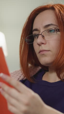 red-haired housewife in glasses reads interesting book sitting on sofa against bright lamp. focused lady flips pages of red textbook at home closeup