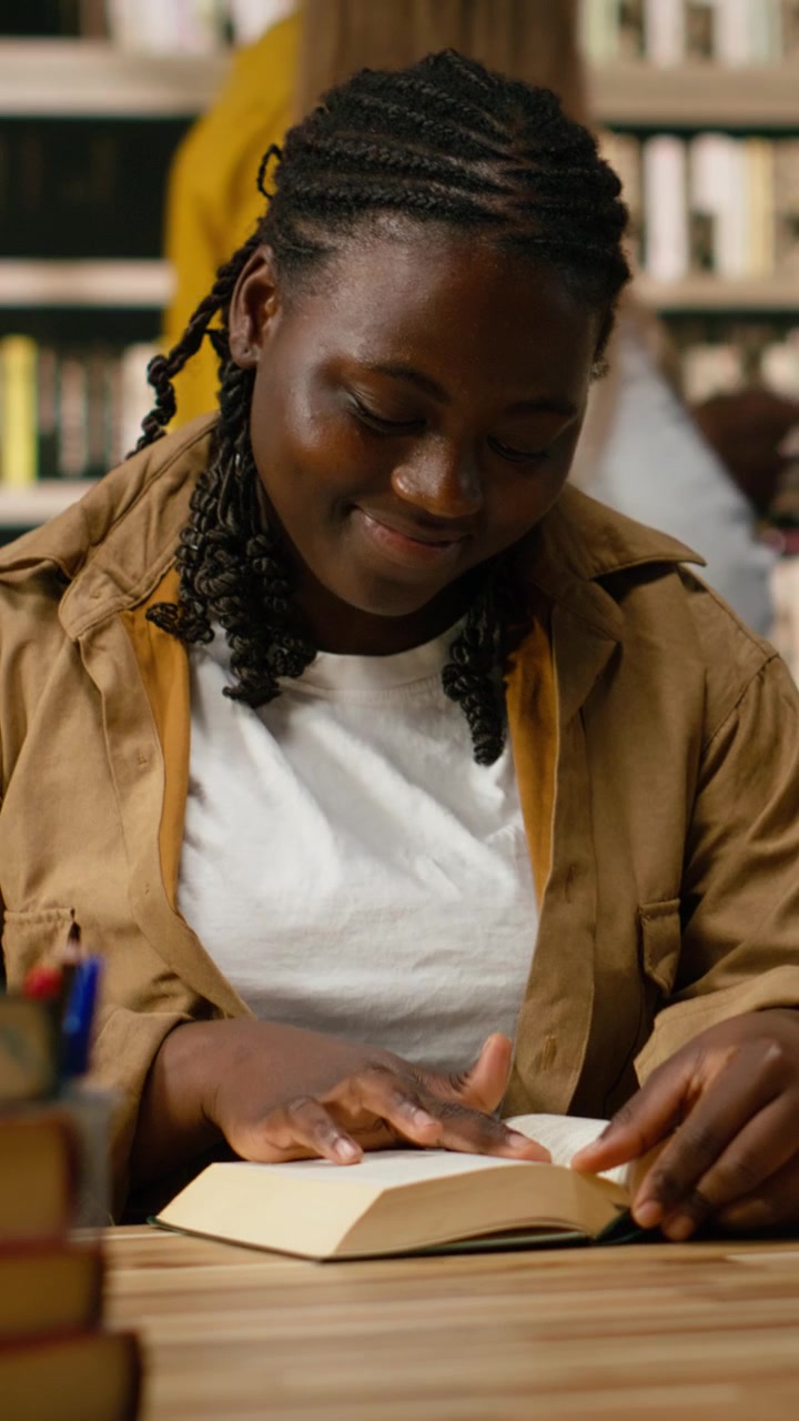 Vertikal video: Afroamerikansk ung voksen åpner en bok og leser på en arbeidsplass på biblioteket