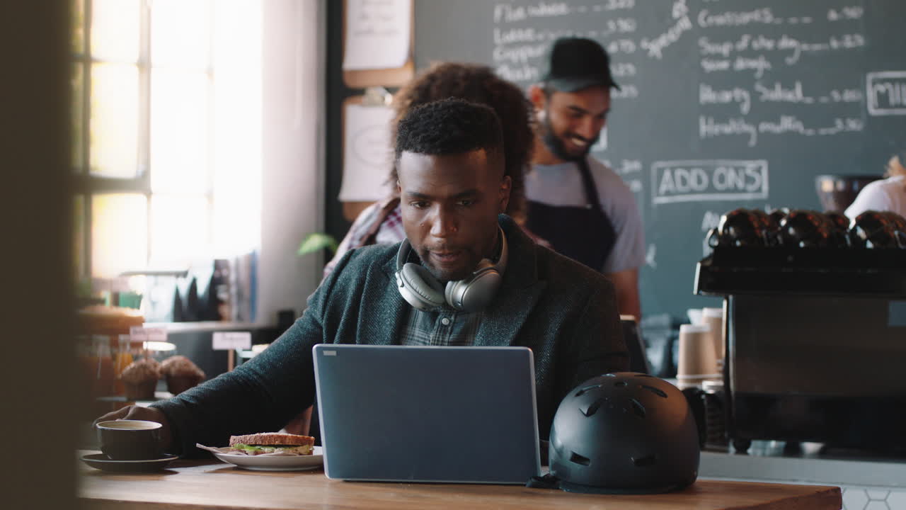giovane afroamericano che usa il portatile in un caffè, naviga online, beve caffè, ascolta musica, indossa le cuffie, gode della tecnologia dei computer mobili