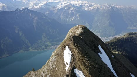 Hombre-Caminando-En-Una-Hermosa-Cima-Entre-Enormes-Montañas-Y-Sobre-Un-Valle-Verde-Con-Un-Lago-Azul