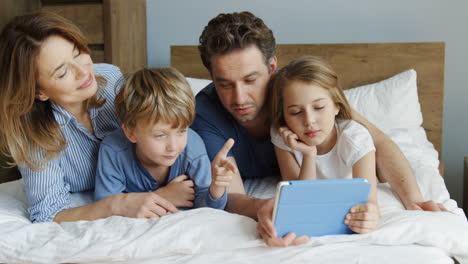 Feliz-Familia-Sonriente-De-Madre,-Padre,-Hija-E-Hijo-Acostados-En-La-Cama-Por-La-Mañana-Y-Haciendo-Videollamadas-En-Una-Tableta