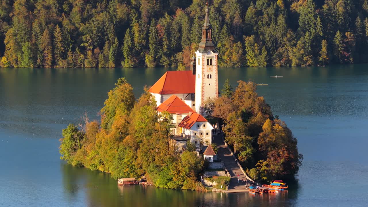 Średni kąt widzenia z lotu ptaka na kościół na wyspie Bled z odbiciami w jeziorze i małymi łodziami wiosłowymi przepływającymi poniżej, blask złotej godziny