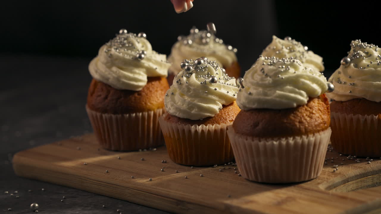 Rows of frosted cupcakes showcased on a tray for serving and enjoyment as hand drops candy pearls on frosting in slow motion