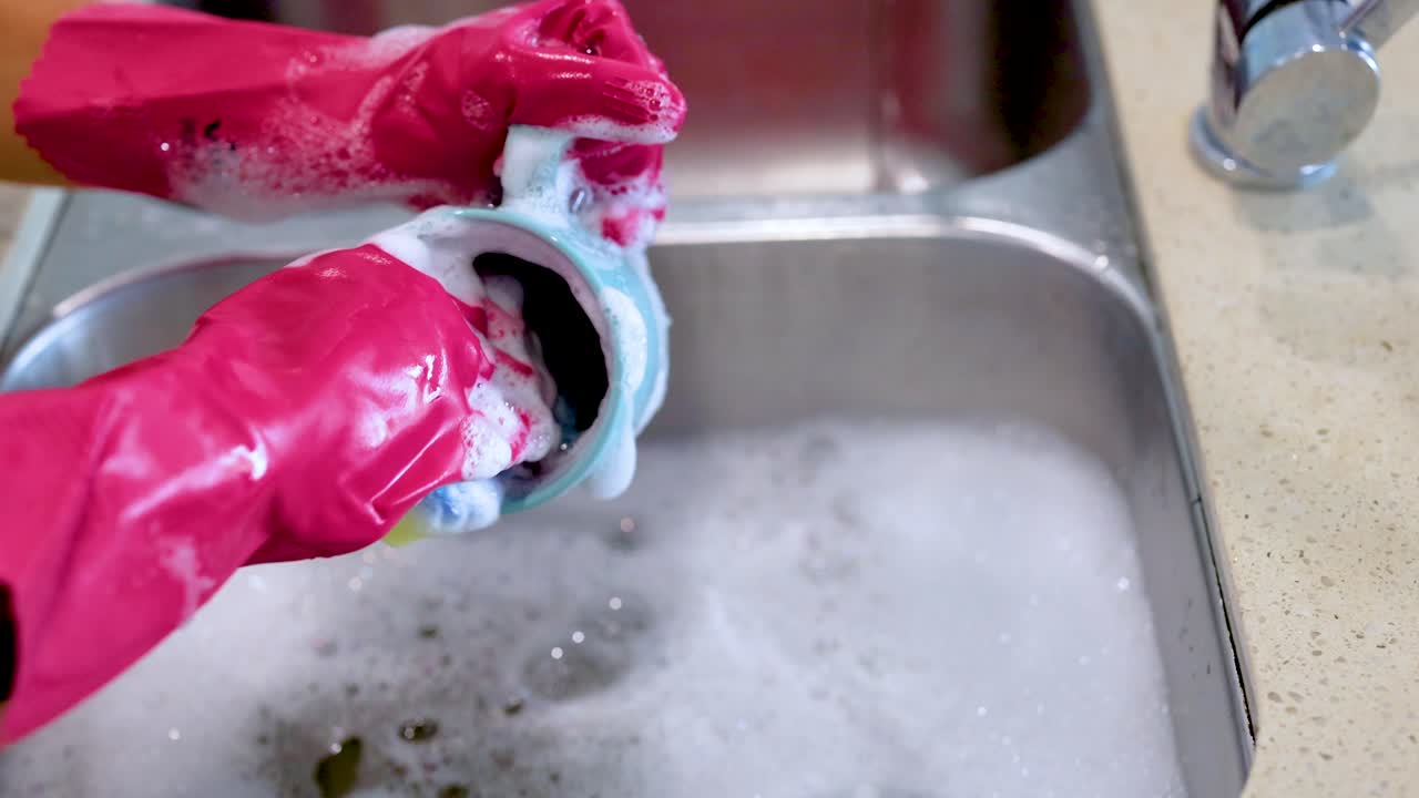 Hands in pink gloves wash a cup in a soapy kitchen sink. Bright lighting highlights the cleaning process