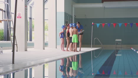 female coach giving children in swimming class briefing as they stand on edge of indoor pool