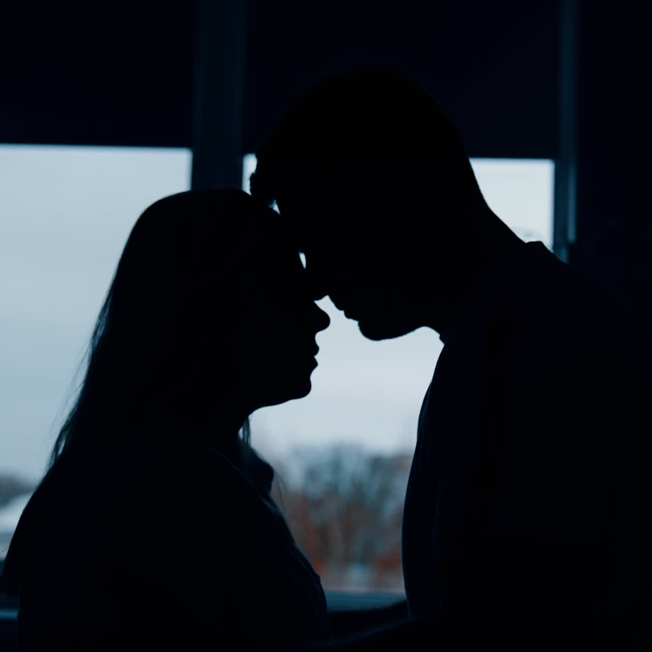 Silhouettes of a couple in love in a dark room standing in front of window. Man soothing and kissing his woman