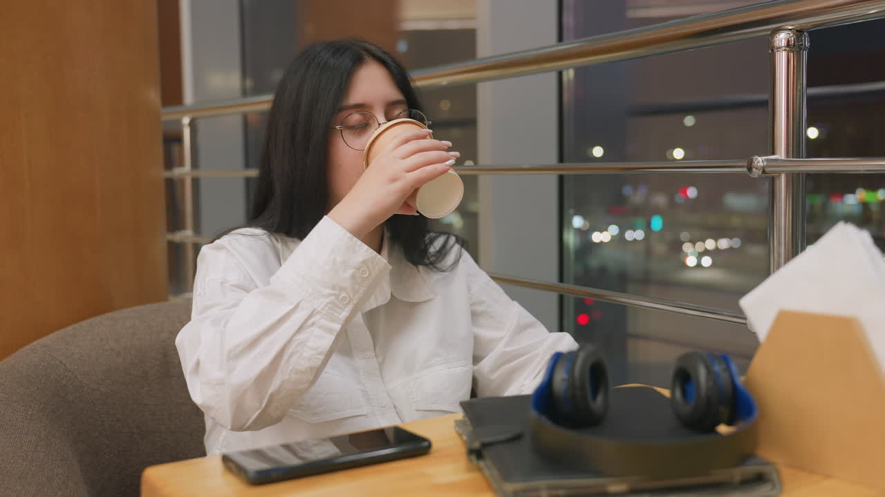 Woman with long black hair in white shirt holds coffee cup and looks outside through glass panel, seated indoors with phone and headphones on table, blurred night city lights visible in background